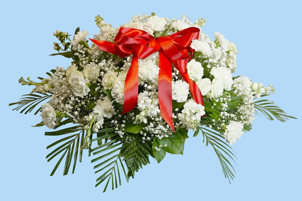A white floral arrangement with green foliage and a large red satin bow, displayed on a white draped surface against a white background.
