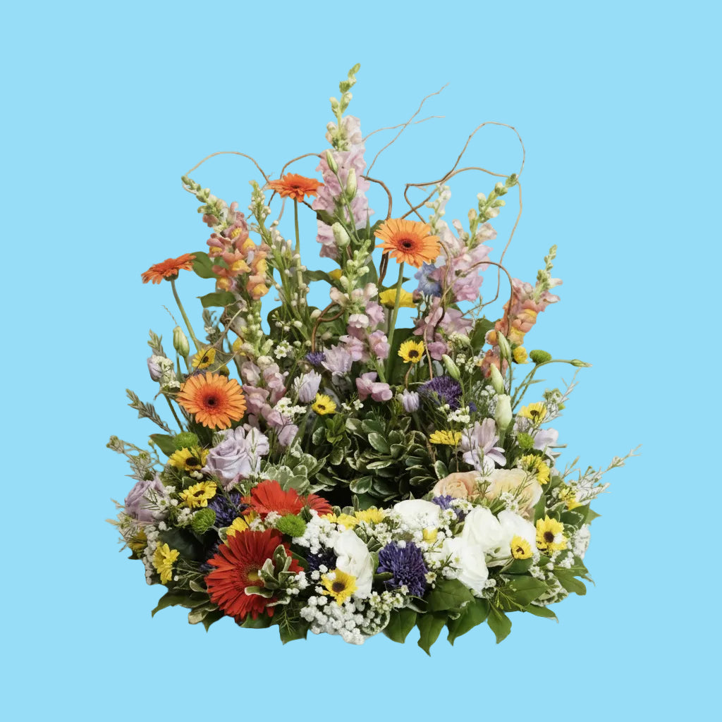 A vibrant, multicolored wildflower arrangement with green foliage, orange, red, yellow, purple, and white blooms, displayed on a white draped studio backdrop.