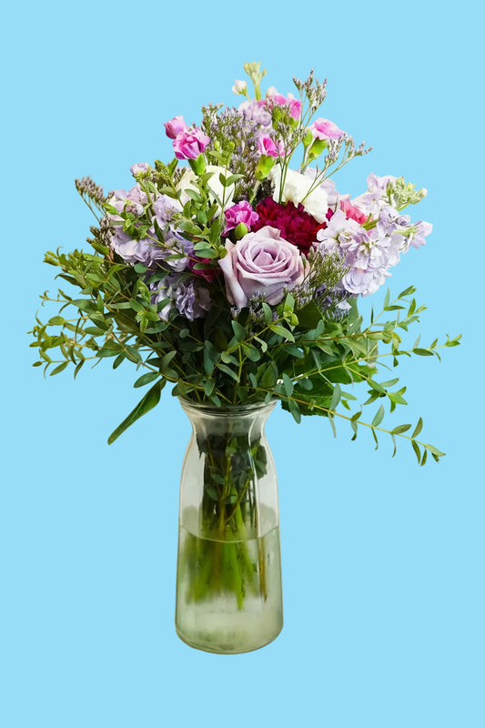A vibrant floral bouquet featuring pink and purple flowers, green foliage, and a light purple rose, arranged in a clear glass vase with water on a white background.