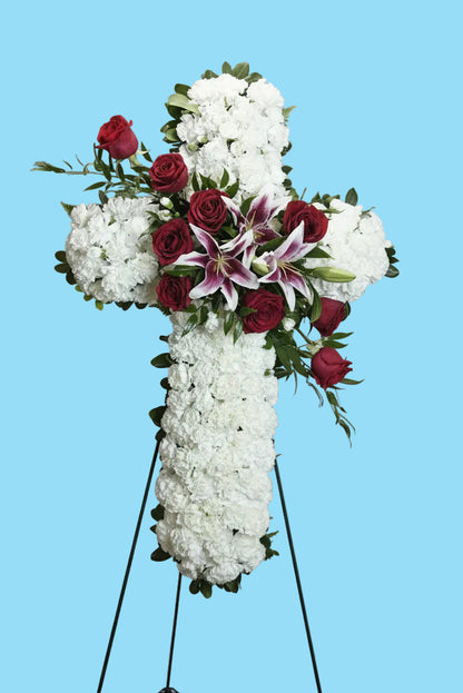 A large floral cross wreath made of white flowers, accented with red roses, pink-and-white lilies, and green foliage, displayed on a black metal stand against a white studio backdrop.