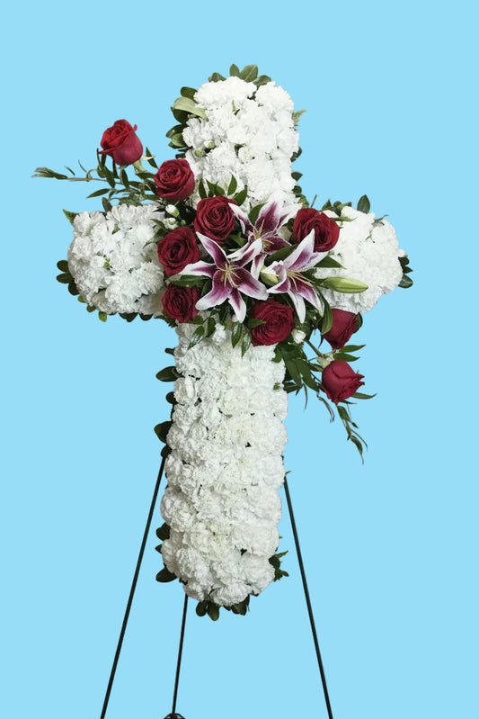 A large floral cross wreath made of white flowers, accented with red roses, pink-and-white lilies, and green foliage, displayed on a black metal stand against a white studio backdrop.