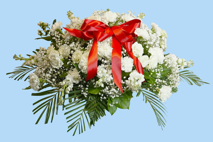 A white floral arrangement with green foliage and a large red satin bow, displayed on a white draped surface against a white background.